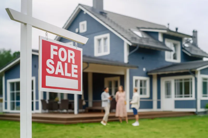 A red For Sale sign is displayed in front of a modern blue house. Three people are standing and talking on the porch in the background. The scene suggests a house showing or real estate transaction.